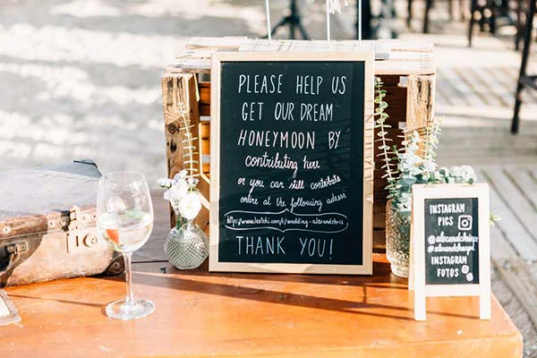 Urne de mariage bohème avec ardoise, fleurs blanches et verre de vin sur une table en bois ensoleillée.