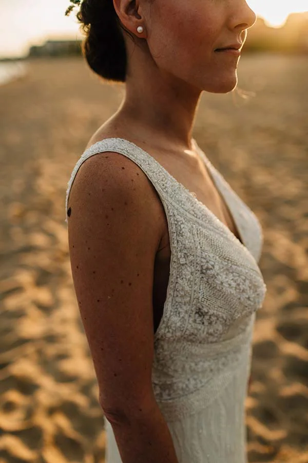 Profil d'une mariée en robe bohème blanche à dentelle, posant sur une plage au coucher du soleil.