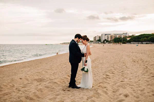Jeunes mariés en tenue chic s'embrassant tendrement sur une plage de sable doré à Barcelone sous un ciel doux.