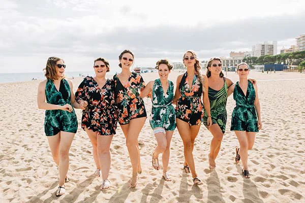 Sept femmes souriantes en robes courtes tropicales vertes et noires marchent ensemble sur une plage ensoleillée.