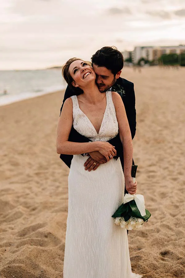 Couple marié complice s'enlaçant sur une plage de Barcelone, elle en robe blanche et lui en costume sombre.
