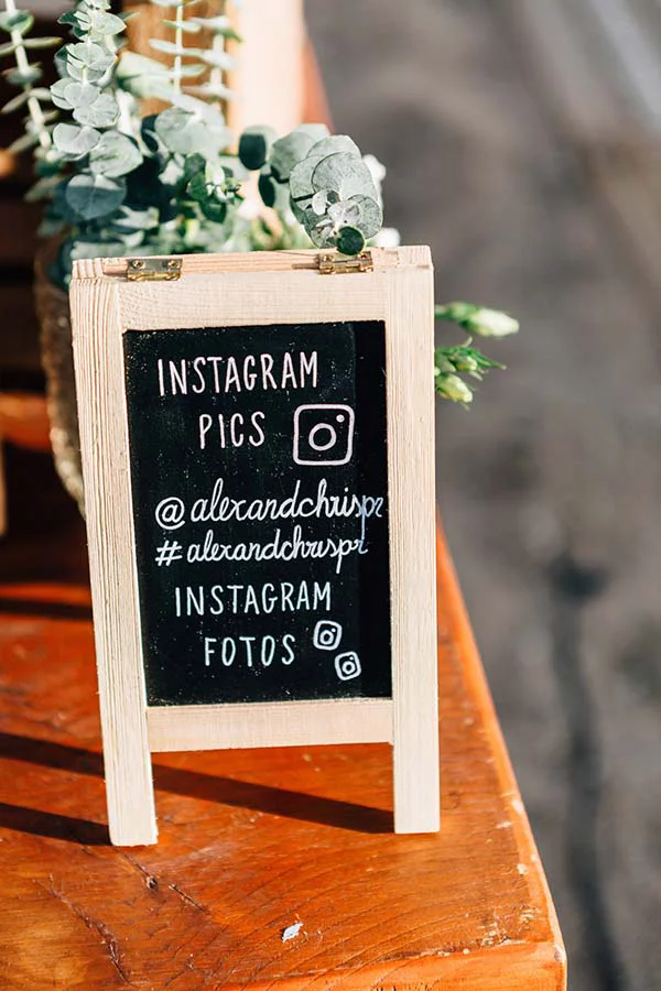 Petit panneau noir en bois clair indiquant le hashtag Instagram du mariage, posé sur une table rustique avec du feuillage.