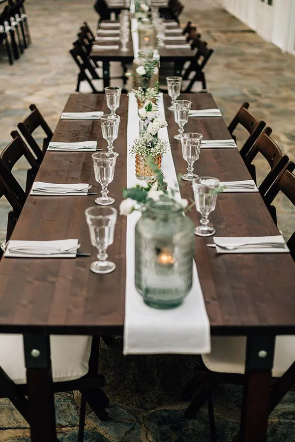 Longue table en bois sombre dressée avec un chemin de table blanc, des fleurs délicates et des verres élégants.