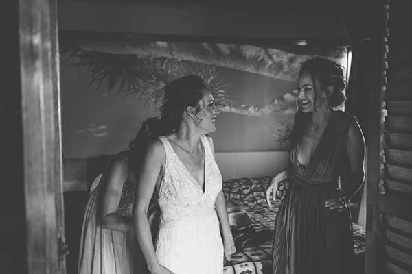 Une mariée et sa demoiselle d'honneur rient ensemble dans une chambre, cliché en noir et blanc à l'ambiance intime.