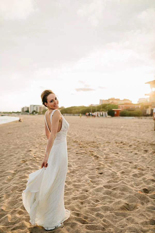 Une mariée souriante en robe blanche fluide pose sur une plage ensoleillée à Barcelone dans une ambiance bohème.