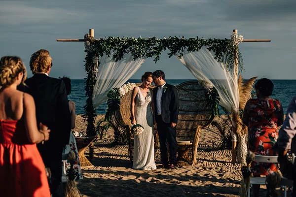 Couple de mariés sous une arche bohème fleurie sur une plage à Barcelone, ambiance chic et romantique au coucher du soleil.