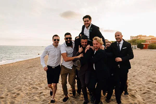 Groupe d'hommes élégants et joyeux s'amusant sur une plage de Barcelone lors d'un mariage chic et bohème.