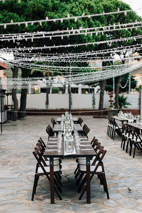 Table de mariage chic et bohème sous des guirlandes lumineuses et des pins, sur une terrasse en pierre à Barcelone.