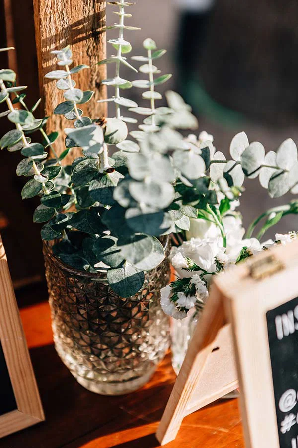 Composition bohème de feuilles d'eucalyptus et fleurs blanches dans un vase en verre ciselé sur un support en bois.