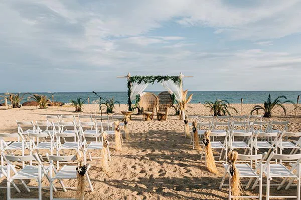 Arche fleurie et chaises blanches sur une plage de sable pour une cérémonie de mariage chic et bohème à Barcelone.
