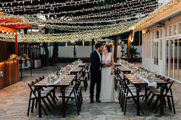 Jeunes mariés s'embrassant lors d'un dîner chic et bohème sous des guirlandes lumineuses à Barcelone.