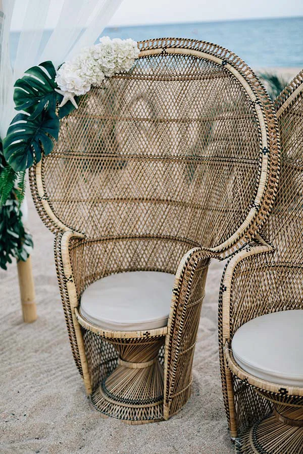 Fauteuils en osier tressé avec coussins blancs et fleurs, installés sur le sable pour un mariage bohème à la plage.