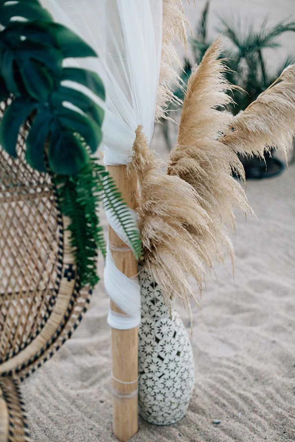 Décoration de mariage bohème sur la plage avec pampas, feuilles tropicales et voilages blancs sur un support en bambou.