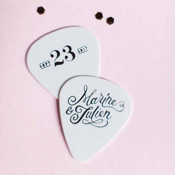 Deux médiators de guitare blancs personnalisés avec les prénoms Marine & Julien et la date du mariage sur fond rose.