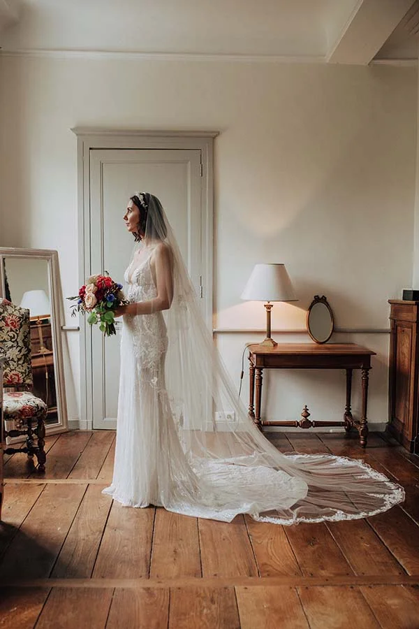 Mariée de profil en robe blanche bohème avec long voile, tenant un bouquet coloré dans un intérieur chaleureux.