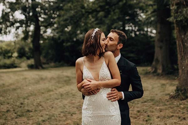 Jeune couple marié s'embrassant tendrement dans un parc boisé, ambiance bohème chic et romantique.