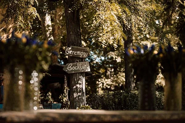 Panneaux en bois indiquant la cérémonie et le cocktail dans un jardin ensoleillé à l'ambiance bohème.