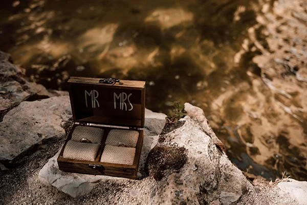 Écrin en bois ouvert avec alliances, marqué Mr & Mrs, posé sur des rochers au bord d'une eau scintillante.