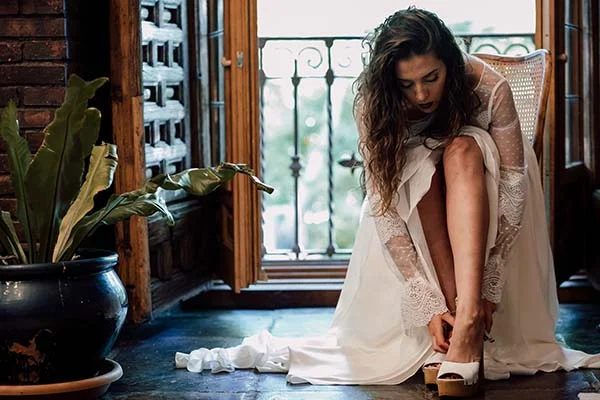 Une mariée en robe blanche bohème ajuste ses chaussures dans un intérieur chaleureux et lumineux à Madrid.
