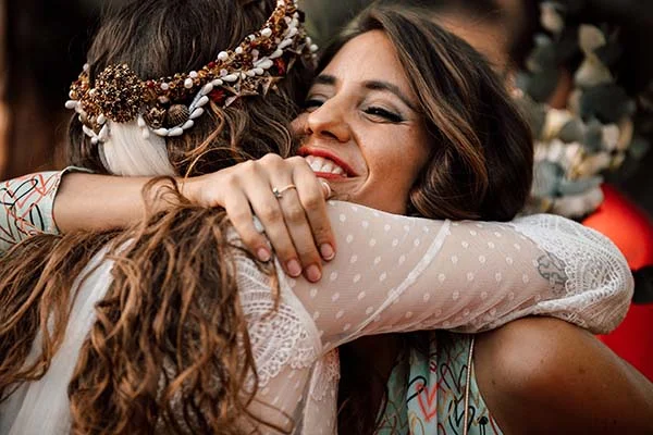 Deux femmes s'enlaçant avec complicité lors d'un mariage bohème, l'une portant une couronne de coquillages.
