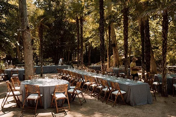 Réception de mariage bohème en plein air sous des palmiers avec des tables nappées de vichy bleu et blanc.