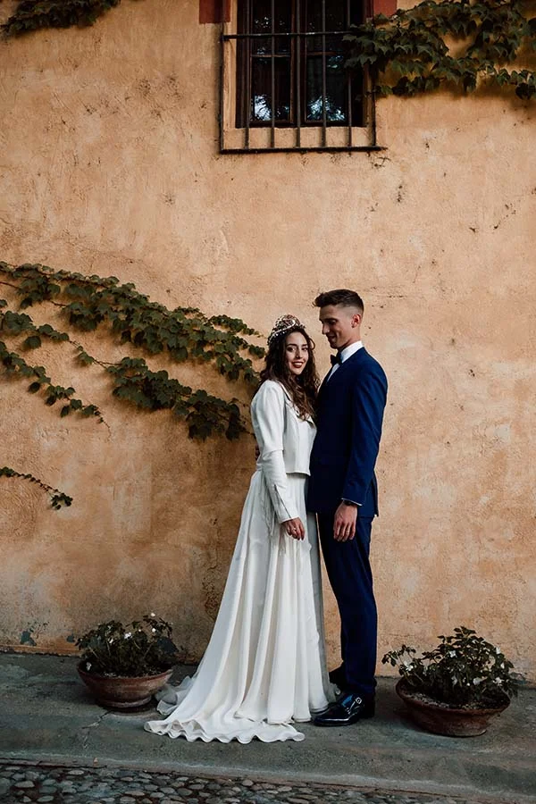 Couple marié élégant posant devant un mur ocre avec des plantes grimpantes dans une ambiance romantique et solaire.