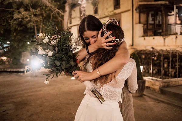 Deux femmes s'enlaçant avec émotion lors d'un mariage bohème nocturne, l'une tenant un bouquet de fleurs blanches.