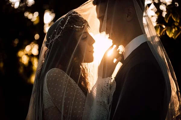 Couple de mariés sous un voile blanc, silhouette à contre-jour au coucher du soleil dans une ambiance bohème et romantique.