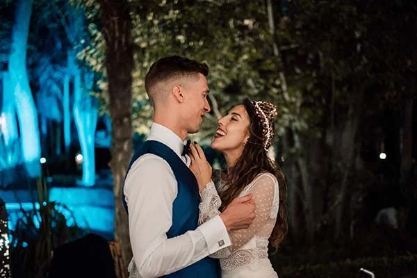 Jeunes mariés complices et souriants dans un jardin nocturne, elle en robe bohème et lui en gilet bleu.