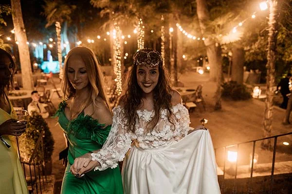 Deux femmes souriantes en robes blanche et verte lors d'une soirée de mariage bohème illuminée par des guirlandes.