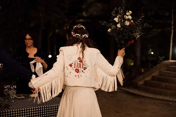 Mariée de dos en veste blanche à franges et couronne de fleurs, tenant un bouquet lors d'un mariage bohème nocturne.