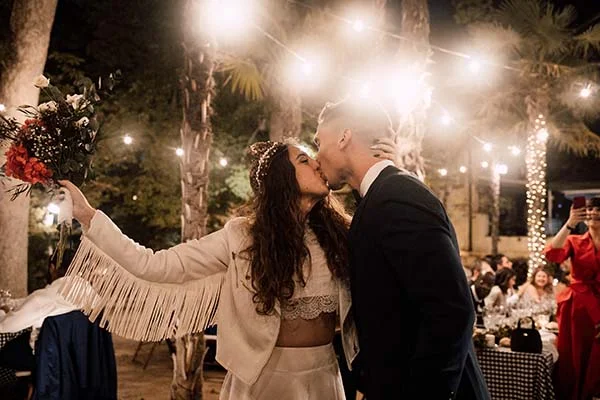 Jeunes mariés s'embrassant sous des guirlandes lumineuses, la mariée portant une veste blanche à franges et un bouquet.