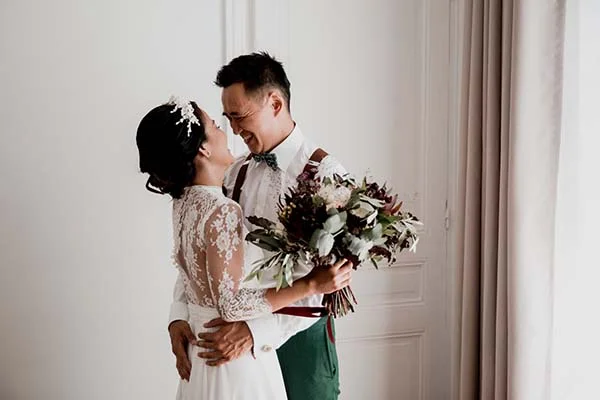 Jeunes mariés complices s'enlaçant, elle en robe blanche dentelée et lui en pantalon vert et nœud papillon.