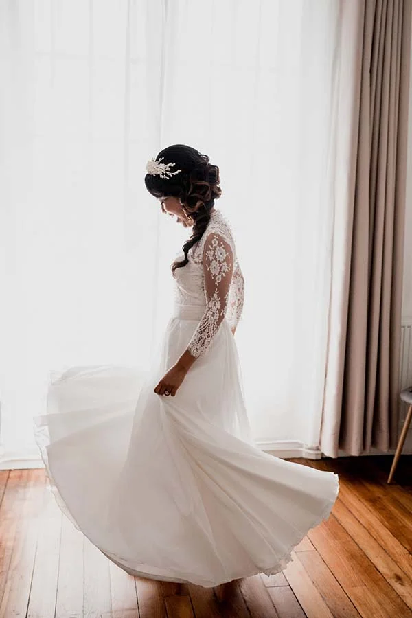 Mariée de profil en robe blanche fluide à manches en dentelle, tournoyant dans une pièce lumineuse et épurée.