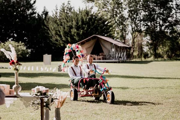 Deux mariés souriants en tenue bohème sur un tricycle rouge dans un jardin avec une tente safari en arrière-plan.