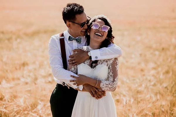 Jeunes mariés riant dans un champ doré, portant des lunettes de soleil et des tenues de mariage au style bohème.
