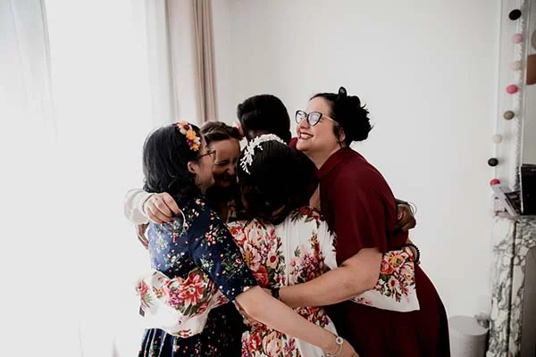 Quatre femmes s'enlacent avec joie, vêtues de robes fleuries et d'une robe bordeaux dans une ambiance chaleureuse.