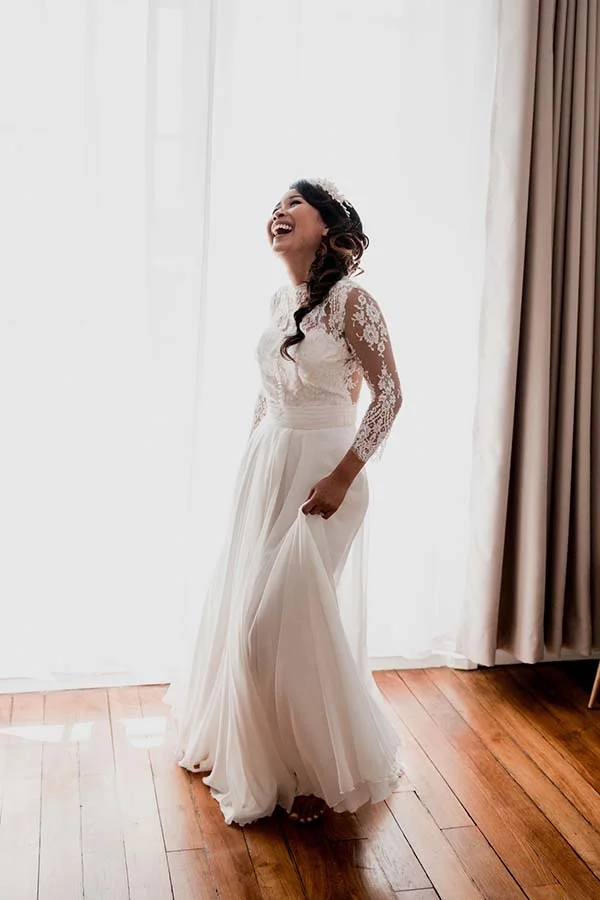Une mariée rayonnante en robe blanche bohème à manches en dentelle, riant devant un rideau lumineux.