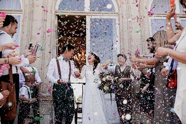 Jeunes mariés riant sous une pluie de confettis colorés, entourés de leurs invités devant un bâtiment blanc.
