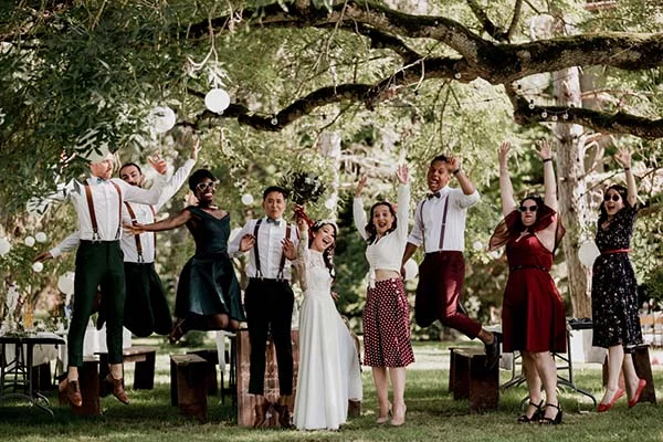 Jeunes mariés et invités sautant de joie sous un grand arbre décoré lors d'un mariage bohème et convivial.