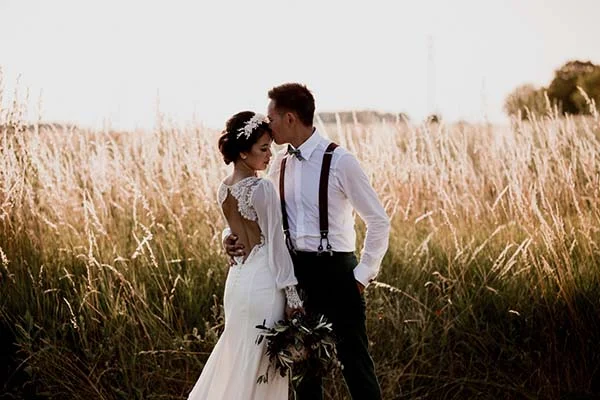 Couple de mariés en tenue bohème s'embrassant tendrement dans un champ d'herbes hautes au coucher du soleil.