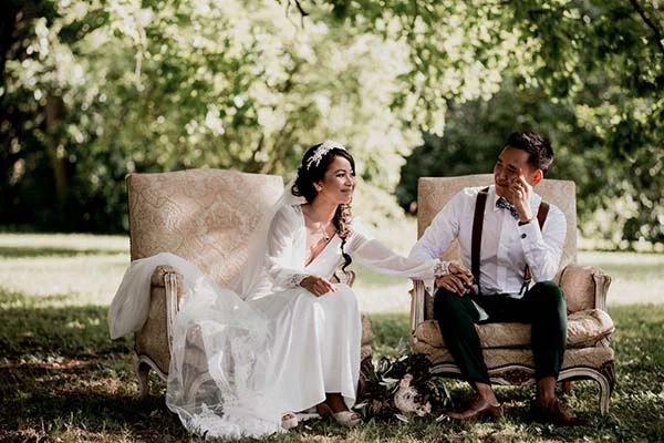 Jeunes mariés en tenue blanche et verte, assis dans des fauteuils vintage en plein air dans une ambiance bohème.