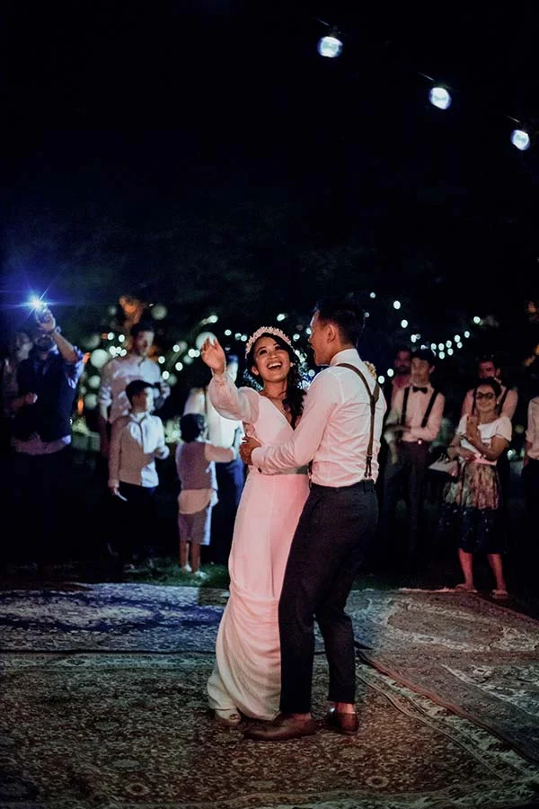 Jeunes mariés dansant avec joie en extérieur la nuit, entourés d'invités sous des guirlandes lumineuses.