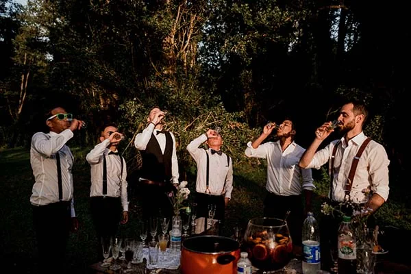 Six hommes en chemises blanches et bretelles trinquant en extérieur dans une ambiance festive et décontractée.
