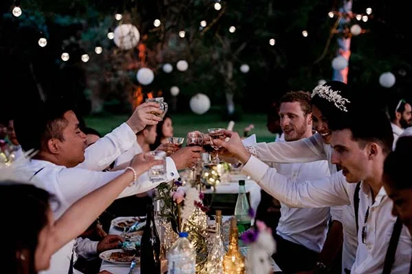 Des invités en chemises blanches portent un toast lors d'un dîner de mariage bohème nocturne sous des guirlandes lumineuses.