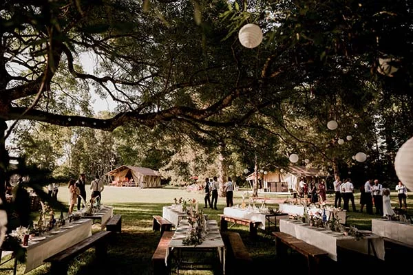 Réception de mariage bohème en plein air avec tables rustiques et lanternes sous de grands arbres ombragés.