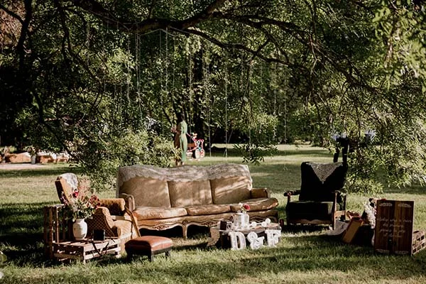 Salon bohème vintage installé sous un grand arbre avec canapé beige, fauteuil noir et décorations artisanales.