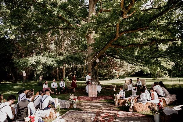 Cérémonie de mariage bohème en plein air sous un grand chêne, avec tapis au sol et invités en tenue claire.