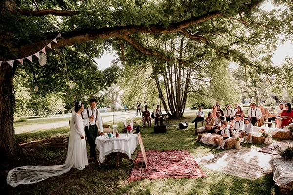 Cérémonie de mariage bohème en plein air sous un grand arbre, avec des invités assis sur des tapis et des coussins.
