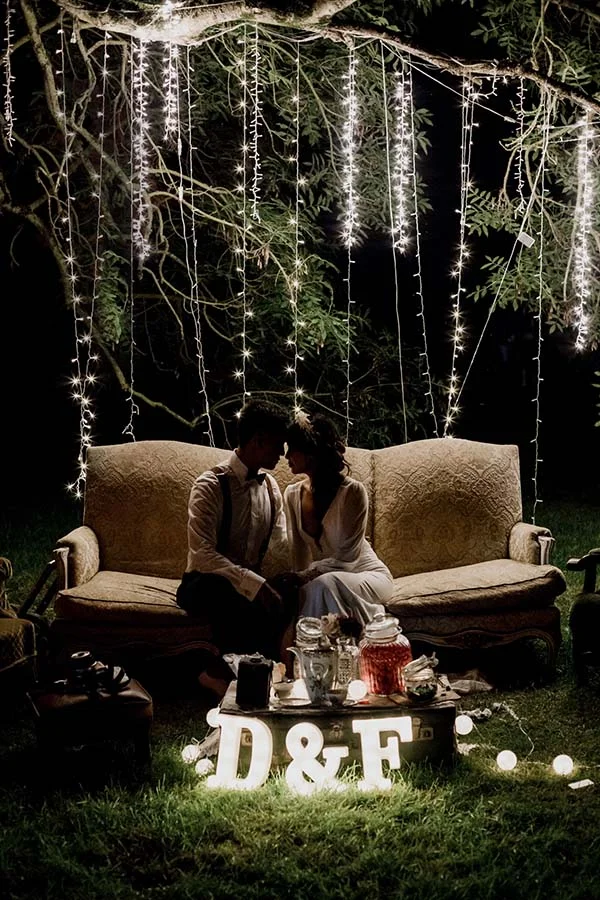 Couple en tenue bohème s'embrassant sur un canapé vintage sous des guirlandes lumineuses nocturnes.